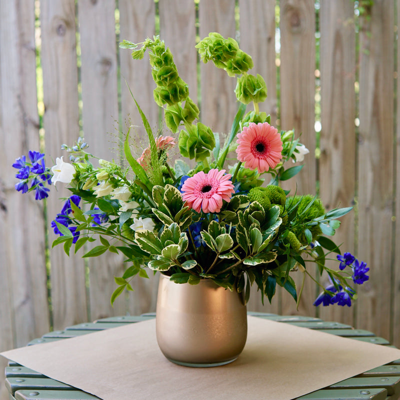 Floral arrangement in a metallic vase on a wooden table with a wooden fence background