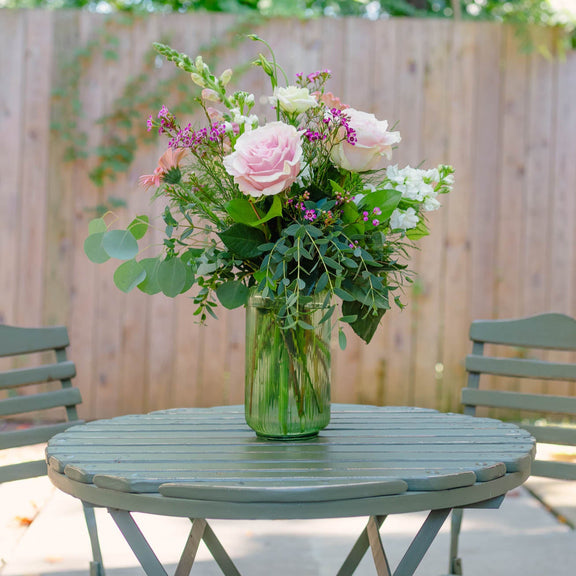 Bouquet of flowers in a vase on a round table outdoors with chairs and greenery.