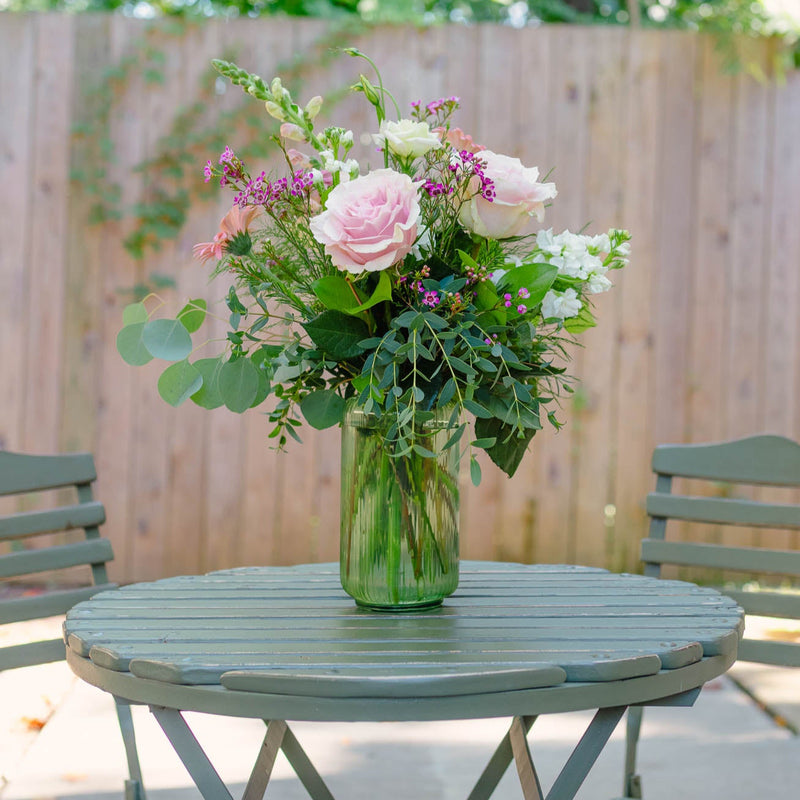 Bouquet of flowers in a vase on a round table outdoors with chairs and greenery.