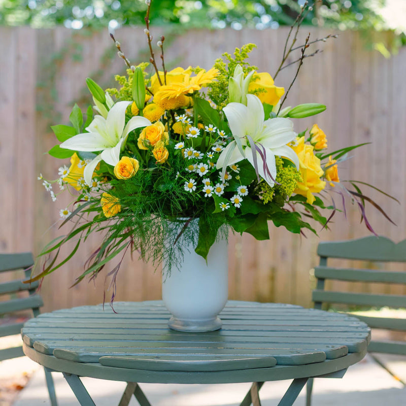 Floral arrangement with yellow and white flowers in a white vase on a green table outdoors.