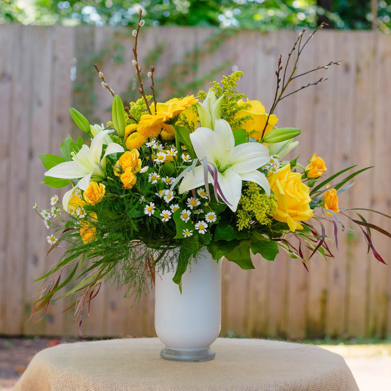 Floral arrangement in a white vase on a table with a burlap tablecloth, outdoors.