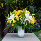 Bouquet of yellow and white flowers in a clear vase on a stone pedestal with greenery in the background