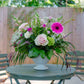 Floral arrangement in a white vase on a round table outdoors with greenery in the background