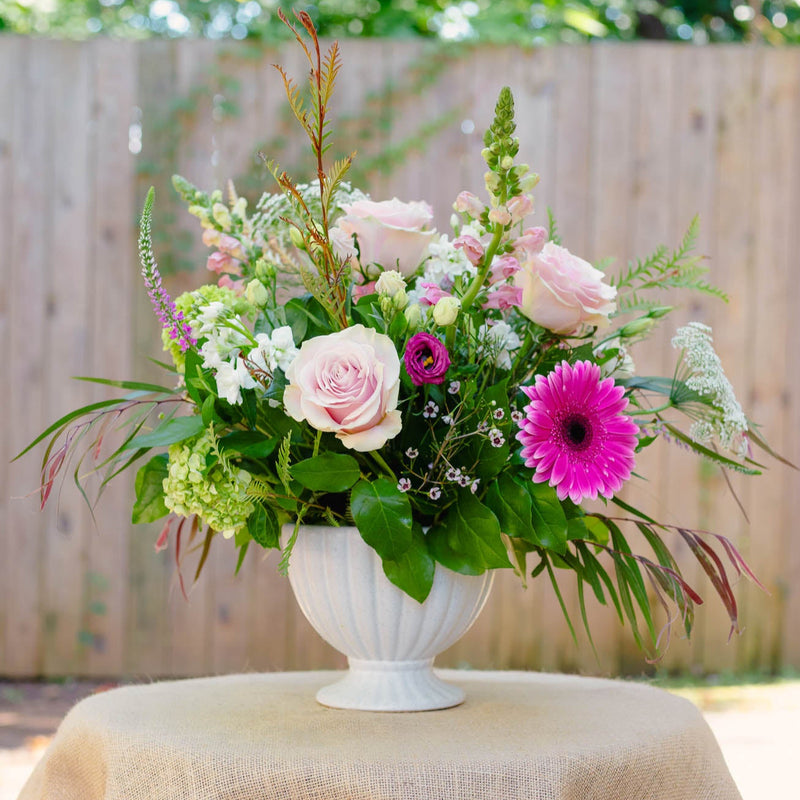 Floral arrangement in a white vase on a table with a burlap tablecloth, outdoors.