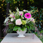 Floral arrangement in a white vase on a stone pedestal with greenery in the background