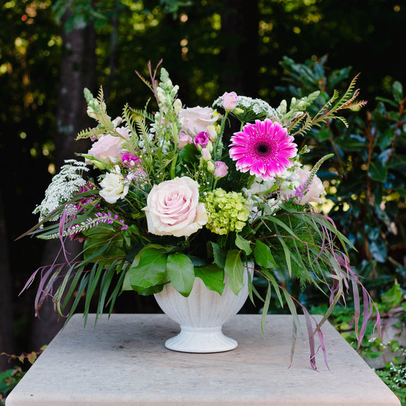 Floral arrangement in a white vase on a stone pedestal with greenery in the background