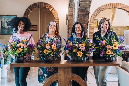 Four women stand behind a table displaying vibrant floral arrangements in various pots, set in a stylish interior space.