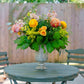 Floral arrangement in a vase on a round table outdoors with chairs and greenery.