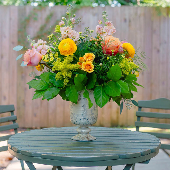 Floral arrangement in a vase on a round table outdoors with chairs and greenery.