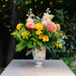 Floral arrangement in a vase on a stone pedestal with greenery in the background
