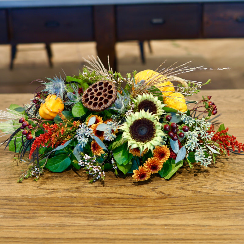 Autumnal flower arrangement with sunflowers, pumpkins, and berries on a wooden surface.