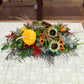 Decorative floral arrangement with pumpkins and sunflowers on a patterned tablecloth.