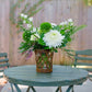 Floral arrangement in a woven basket on a green table with chairs outdoors.