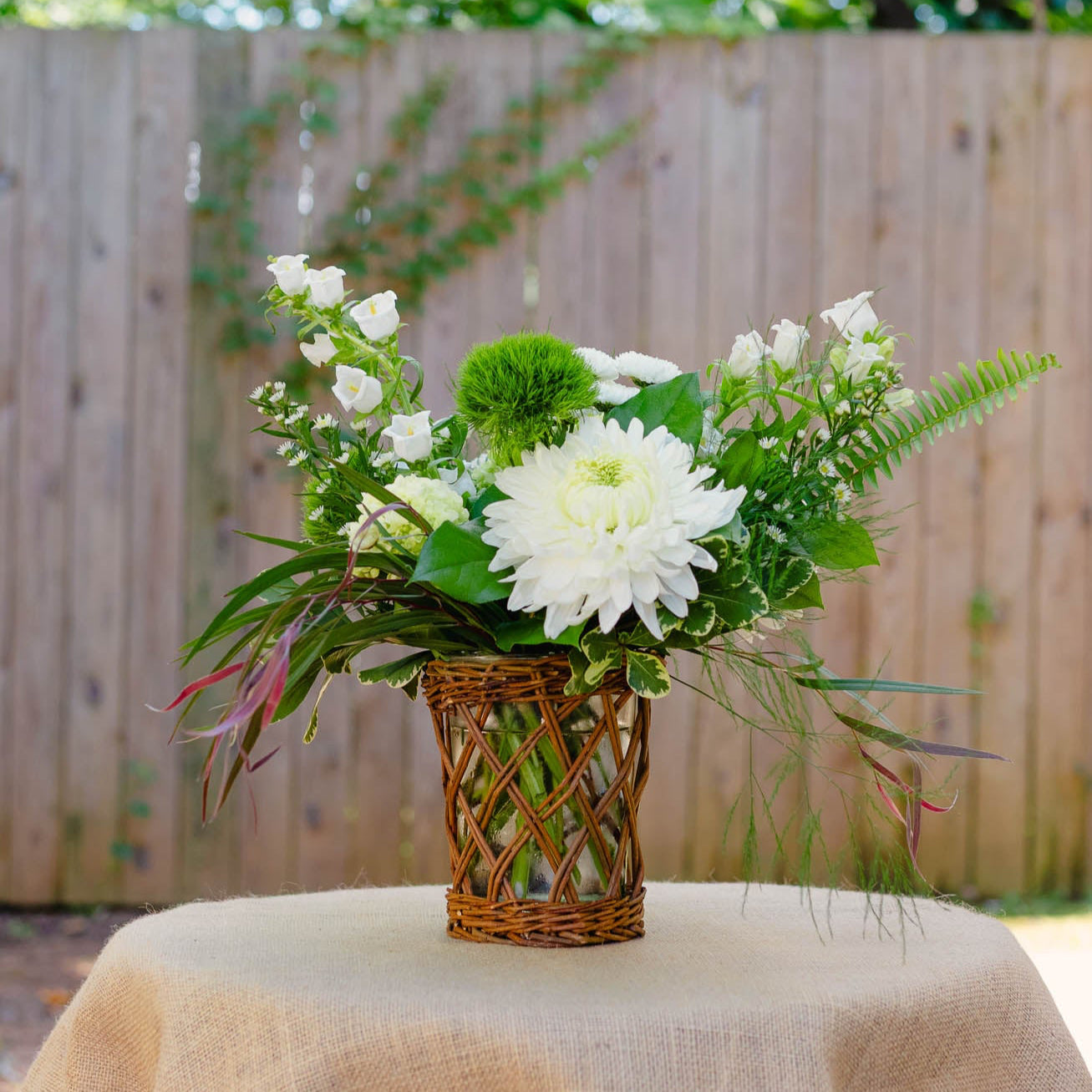 Floral arrangement in a woven basket on a table with a burlap tablecloth, outdoors.