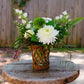 Floral arrangement in a woven vase on a wooden table with a wooden fence and greenery in the background