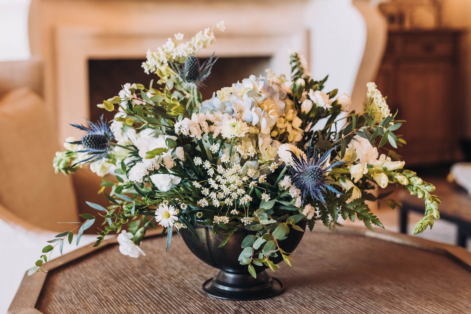 A decorative floral arrangement in a black vase, featuring white and blue flowers, set on a wooden table in a cozy interior.
