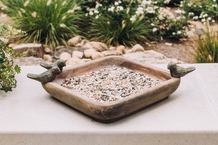 A decorative bird feeder with seeds, featuring two small bird figurines perched on its edges, set against a lush garden background.