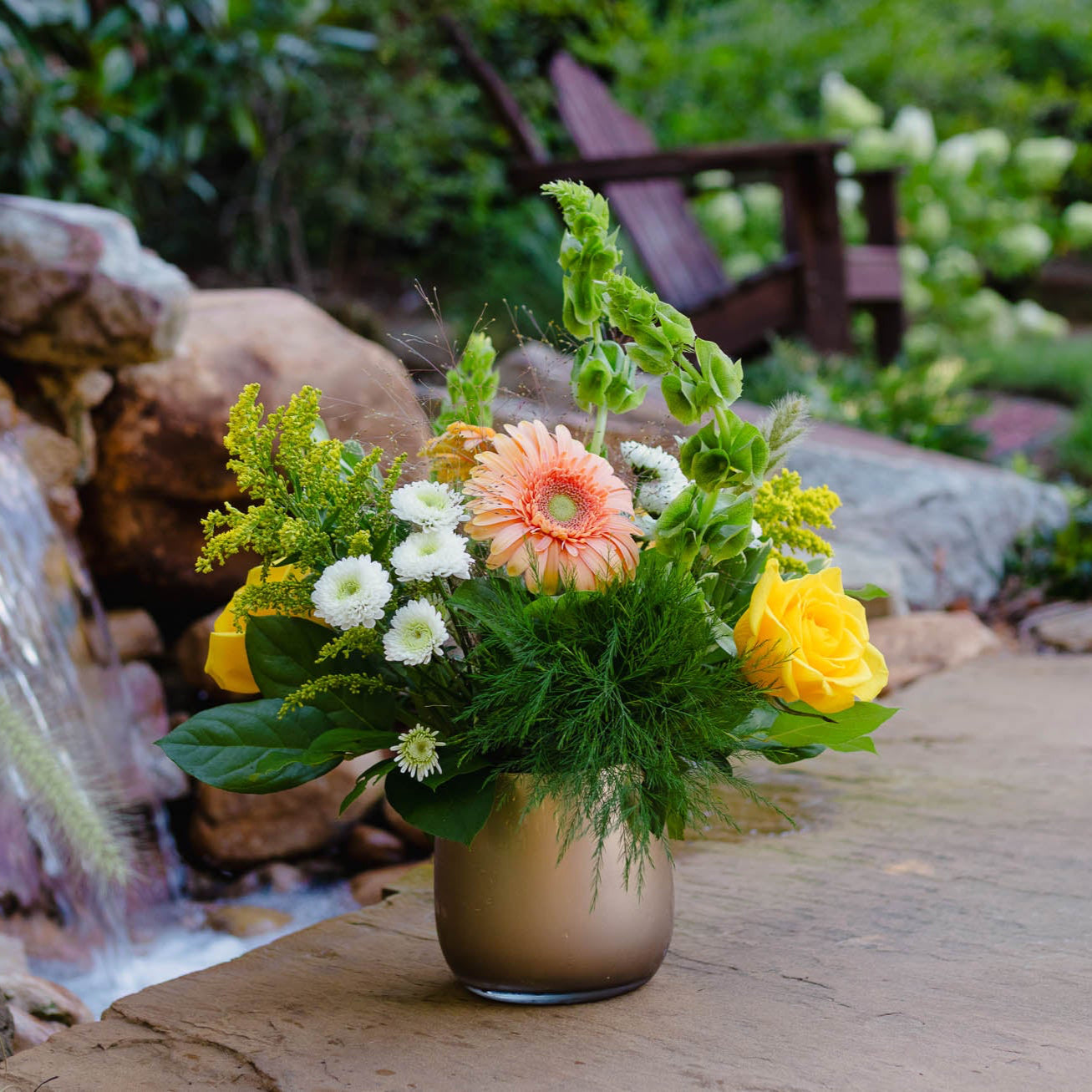 Floral arrangement in a vase on a wooden surface with a water feature in the background
