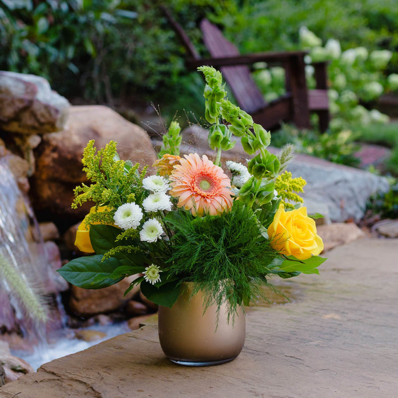 Floral arrangement in a vase on a wooden surface with a water feature in the background