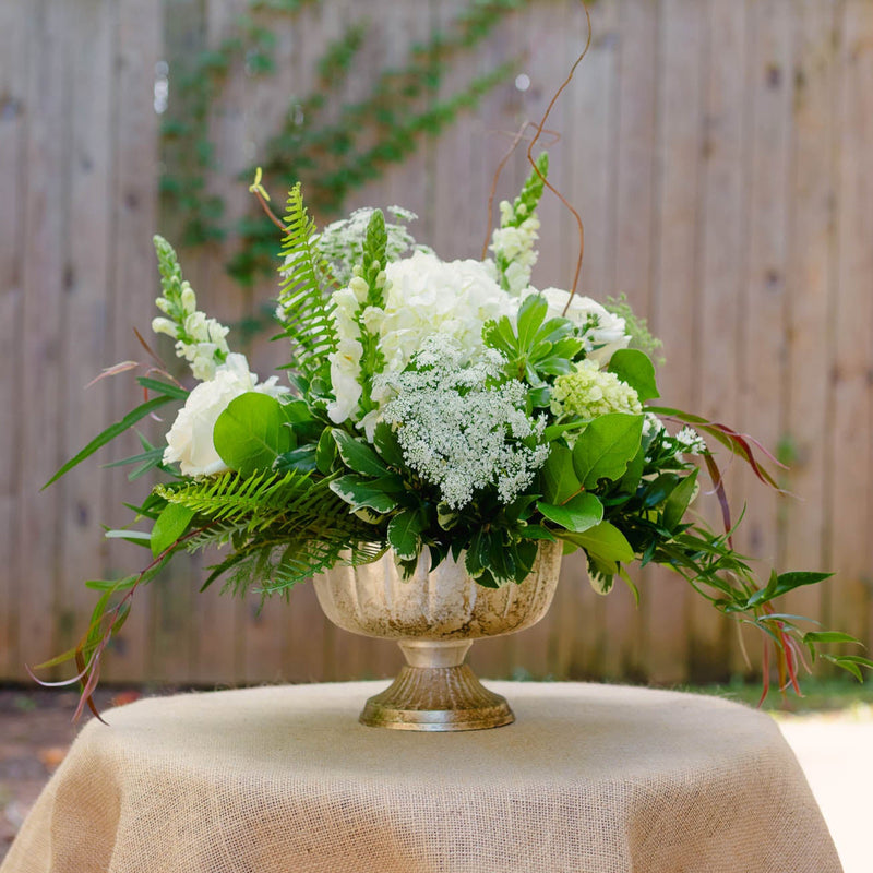 Floral arrangement in a gold vase on a table with a burlap tablecloth, outdoors.