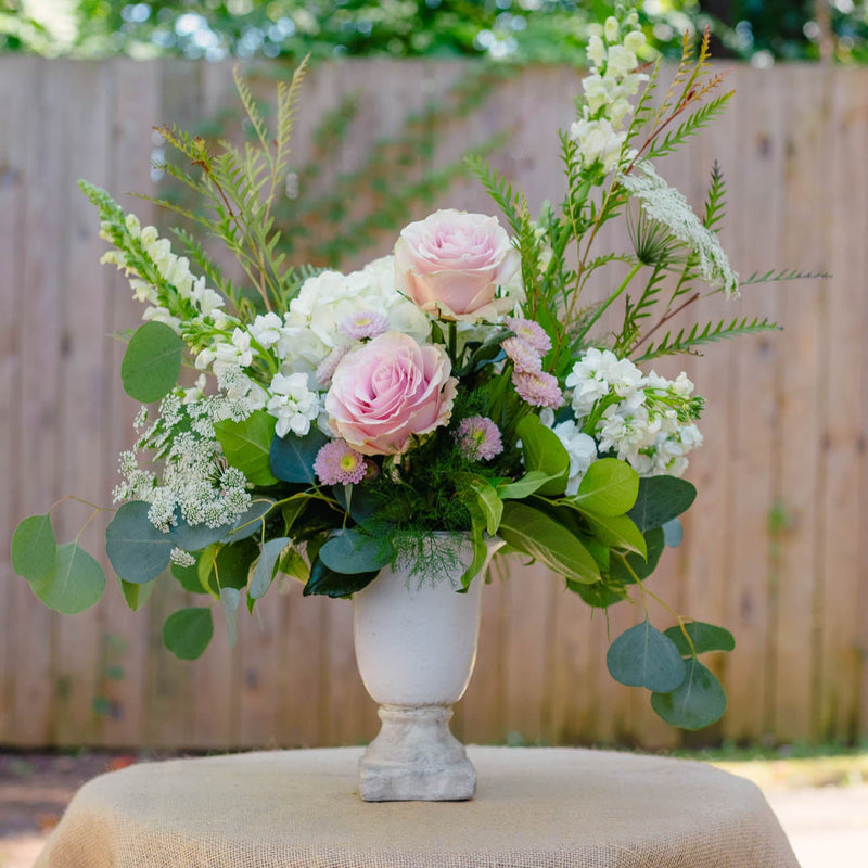 Floral arrangement in a white vase on a table with a burlap tablecloth, outdoors.
