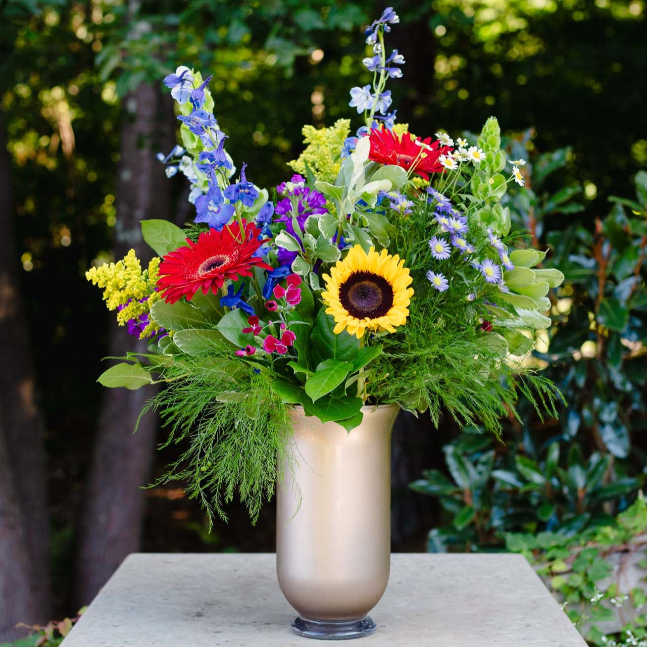 Colorful flower arrangement in a vase on a stone pedestal with greenery in the background