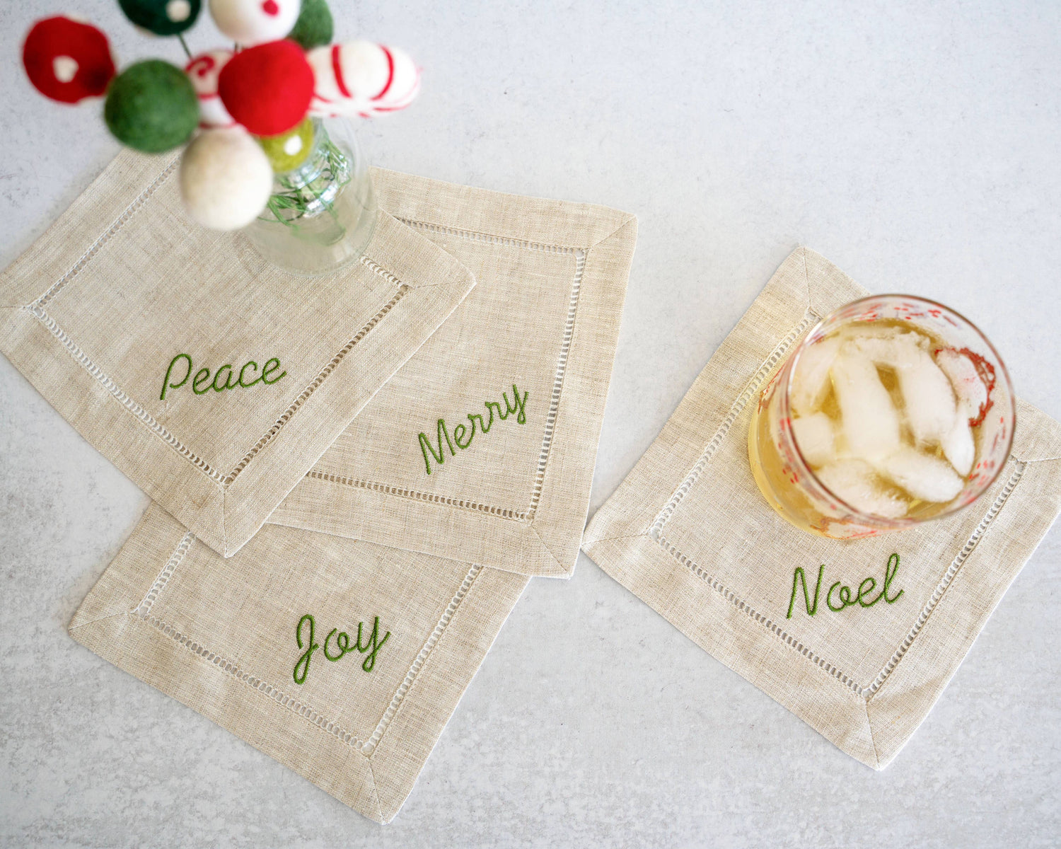 Beige cocktail napkins with Christmas-themed text on a white surface with a glass of iced drink.
