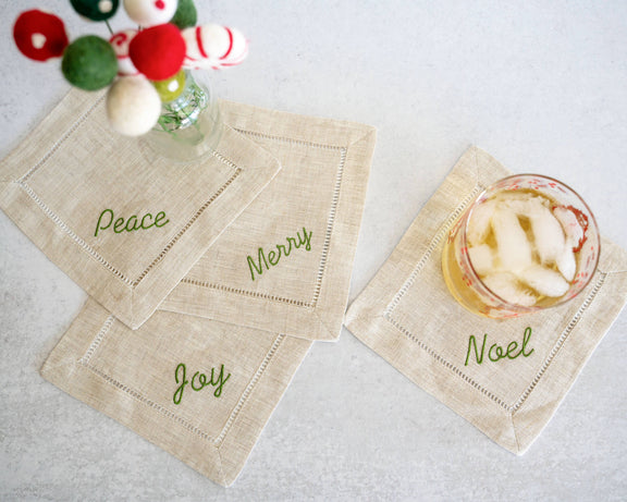 Beige cocktail napkins with Christmas-themed text on a white surface with a glass of iced drink.